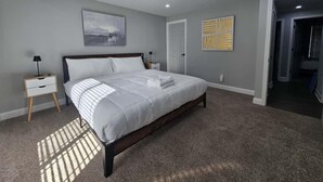 A bedroom with a large bed, a nightstand, a wall-mounted lamp, and a door leading to another room.