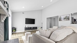 A modern living room with a grey sofa, a flat-screen TV mounted on the wall, a small side table, and a staircase leading to an upper level.