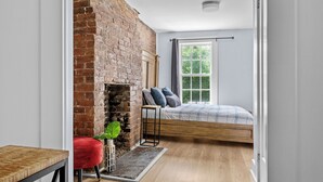 A bedroom with a brick fireplace, a bed with a plaid duvet, a red chair, and a small table. - Historic 2 bedroom central down town location near waterfront - #3 (Halifax)