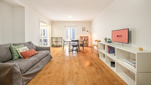 Un salon moderne avec un canapé gris, un plancher en bois, un coin repas avec une table et des chaises, une bibliothèque et un téléviseur affichant le message « PRÊT POUR LES INVITÉS ».