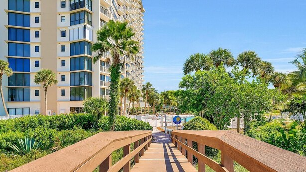 A wooden bridge leading to a high-rise building with balconies, surrounded by lush greenery and palm trees. - Beachfront Condo With Pool, Tennis and Fitness Center (Marco Island)
