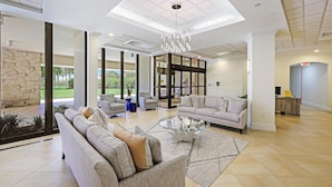 A spacious lobby with a modern chandelier, comfortable seating, and large windows offering a view of the outdoors. - Beachfront Condo With Pool, Tennis and Fitness Center (Marco Island)