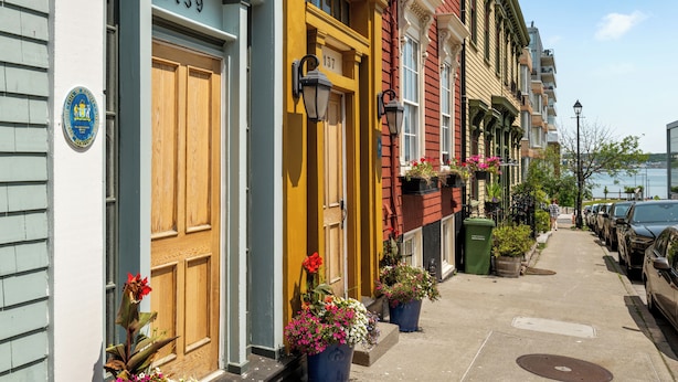 A row of colorful townhouses with numbered doors, a sidewalk, parked cars, and a body of water in the background.