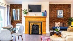 A living room with a fireplace, a television mounted above it, and a brick wall. There are chairs, a table, and a sofa.