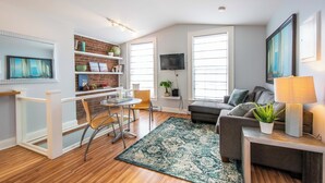 A modern living room with a dining area, a grey sofa, a TV mounted on the wall, and a bookshelf with decorative items. - Historic 1BR DT in amazing location steps from waterfront in Old South District (Halifax)