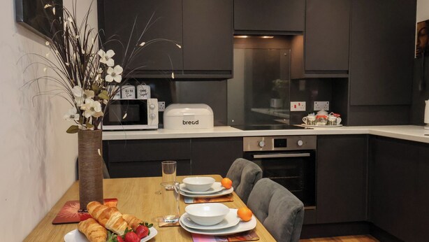 A modern kitchen with dark cabinets, a white countertop, and a dining table set with croissants, strawberries, and oranges. - The Arches (Whitby)