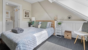 Ein Schlafzimmer mit geneigter Decke, einem Bett mit gesteppter Bettdecke, einem Nachttisch mit Lampe, einem Sessel und einer kleinen Pflanze.