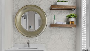 A bathroom with a white vanity, a round mirror, a toilet, and a shelf with plants.