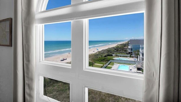 Une vue à travers une fenêtre donnant sur une plage et des bâtiments situés au bord de la piscine.