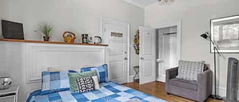 A bedroom with a large bed, a grey armchair, a standing heater, and a chandelier.