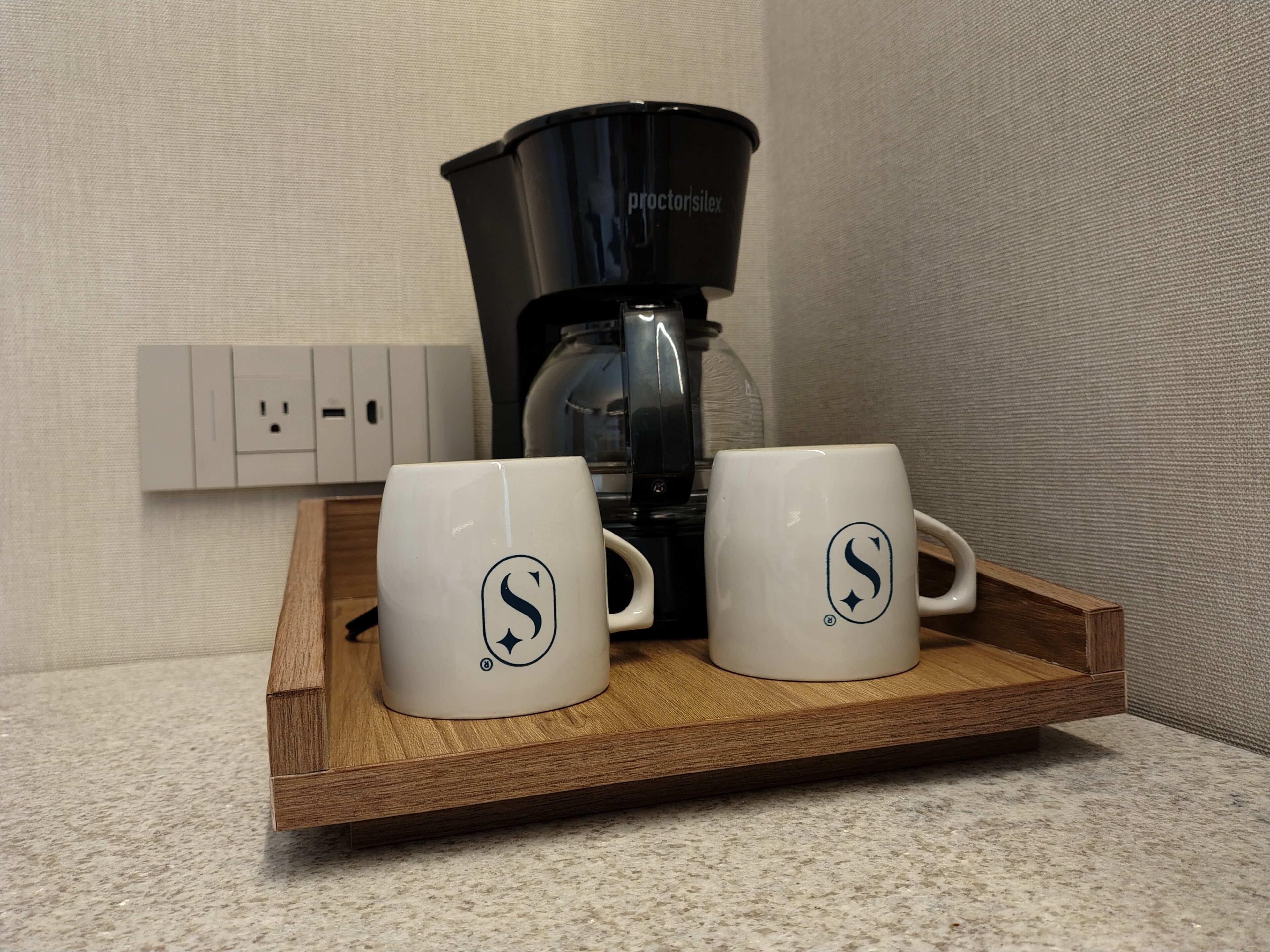 A coffee maker on a wooden tray with two white mugs featuring a logo.