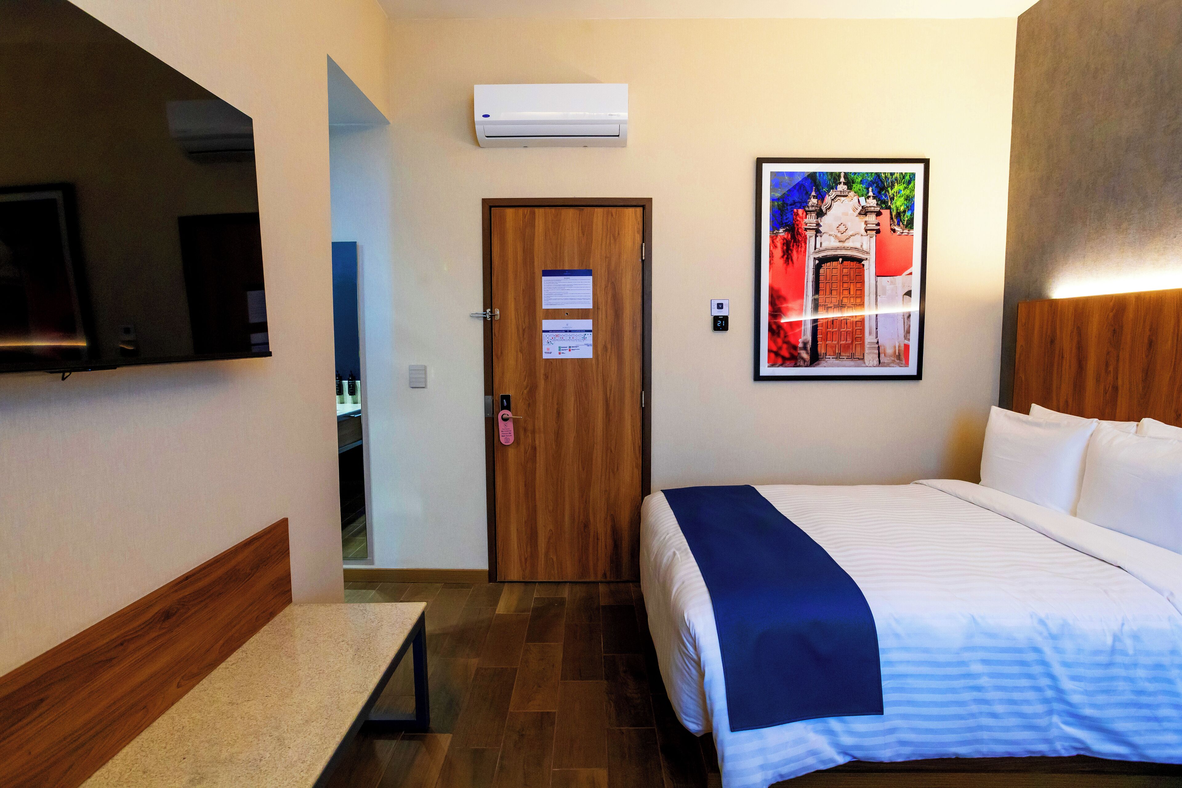 A hotel room with a bed, a bench, a TV, an air conditioner, a door, and a framed picture on the wall.