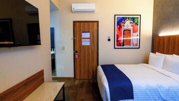 A hotel room with a bed, a bench, a TV, an air conditioner, a door, and a framed picture on the wall.