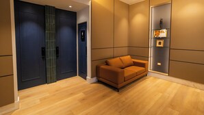 Een moderne hotellobby met een houten vloer, een bruine bank, donkerblauwe deuren en een wandplank met ingekaderde foto's.