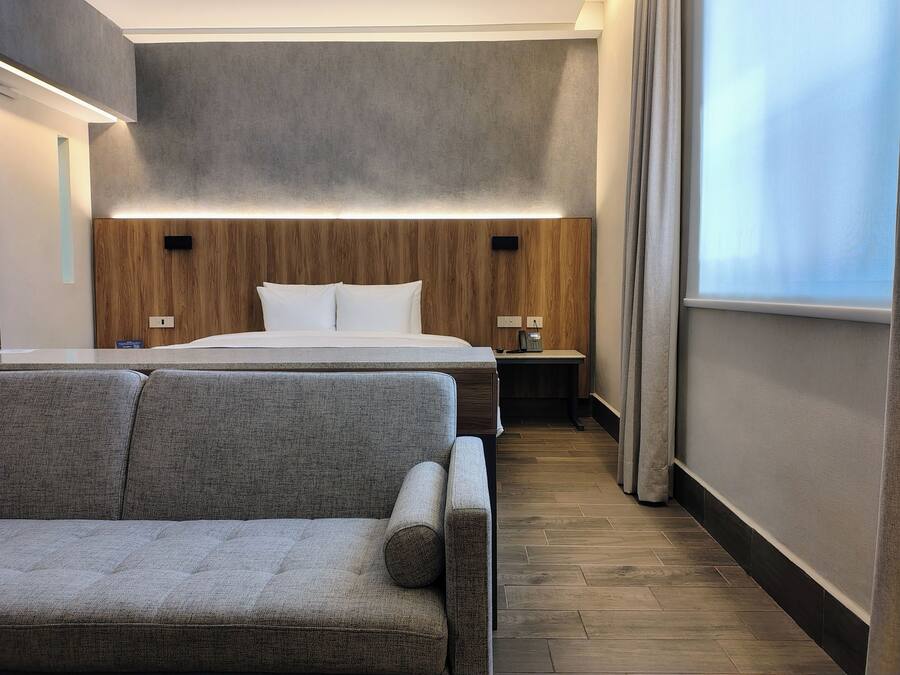Una habitación de hotel con cama, mesita de noche, lámpara, sofá y un amplio ventanal.