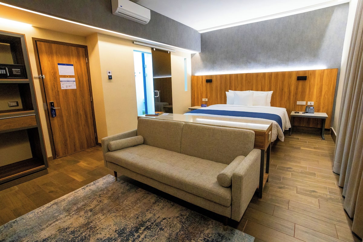 A hotel room with a sofa, a bed with a wooden headboard, a bedside table, and a small desk with a chair.
