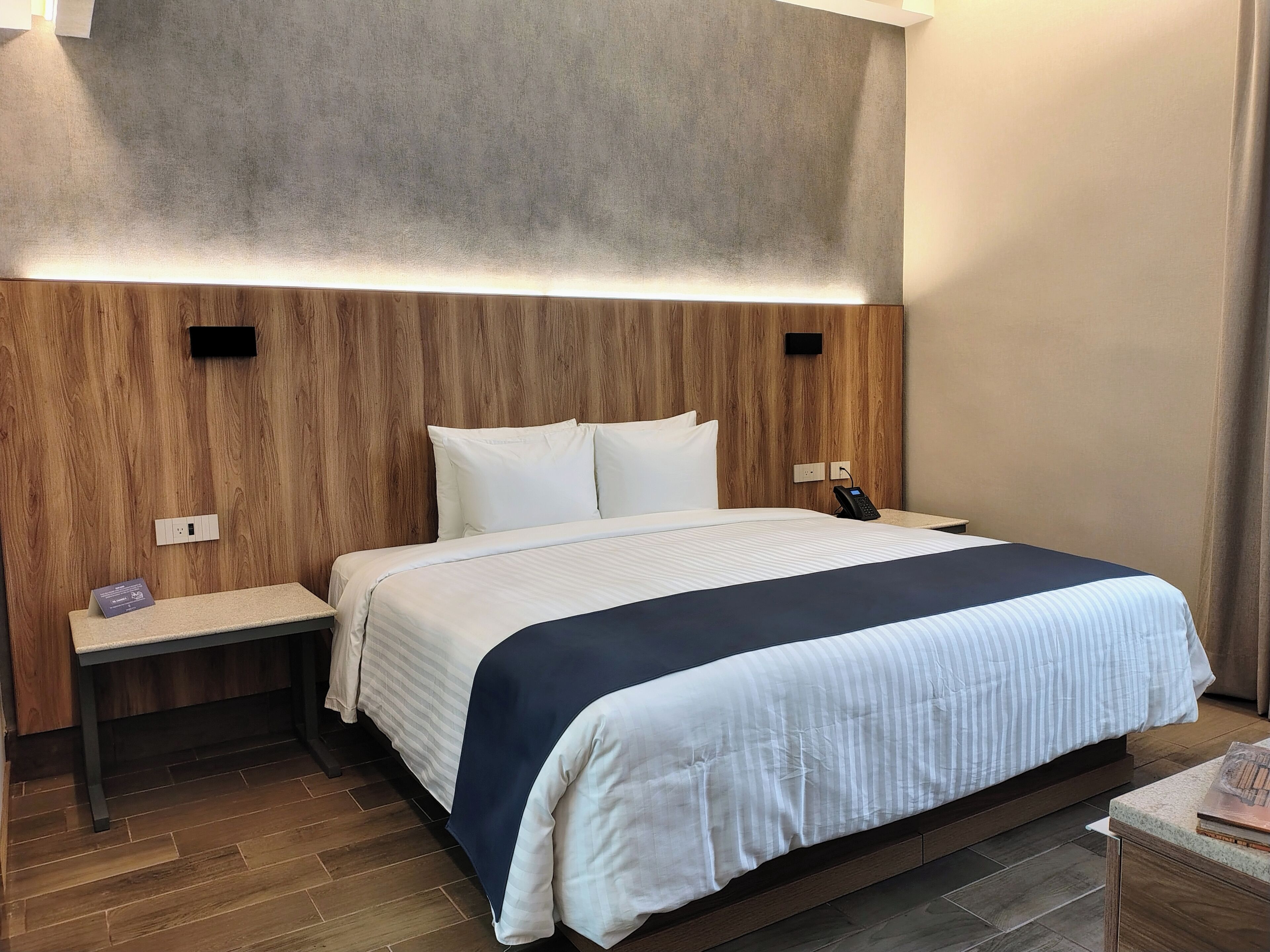 Habitación de hotel con cabecera de madera, cama con ropa de cama blanca y azul marino, mesita de noche con teléfono y una pequeña estantería con un libro.