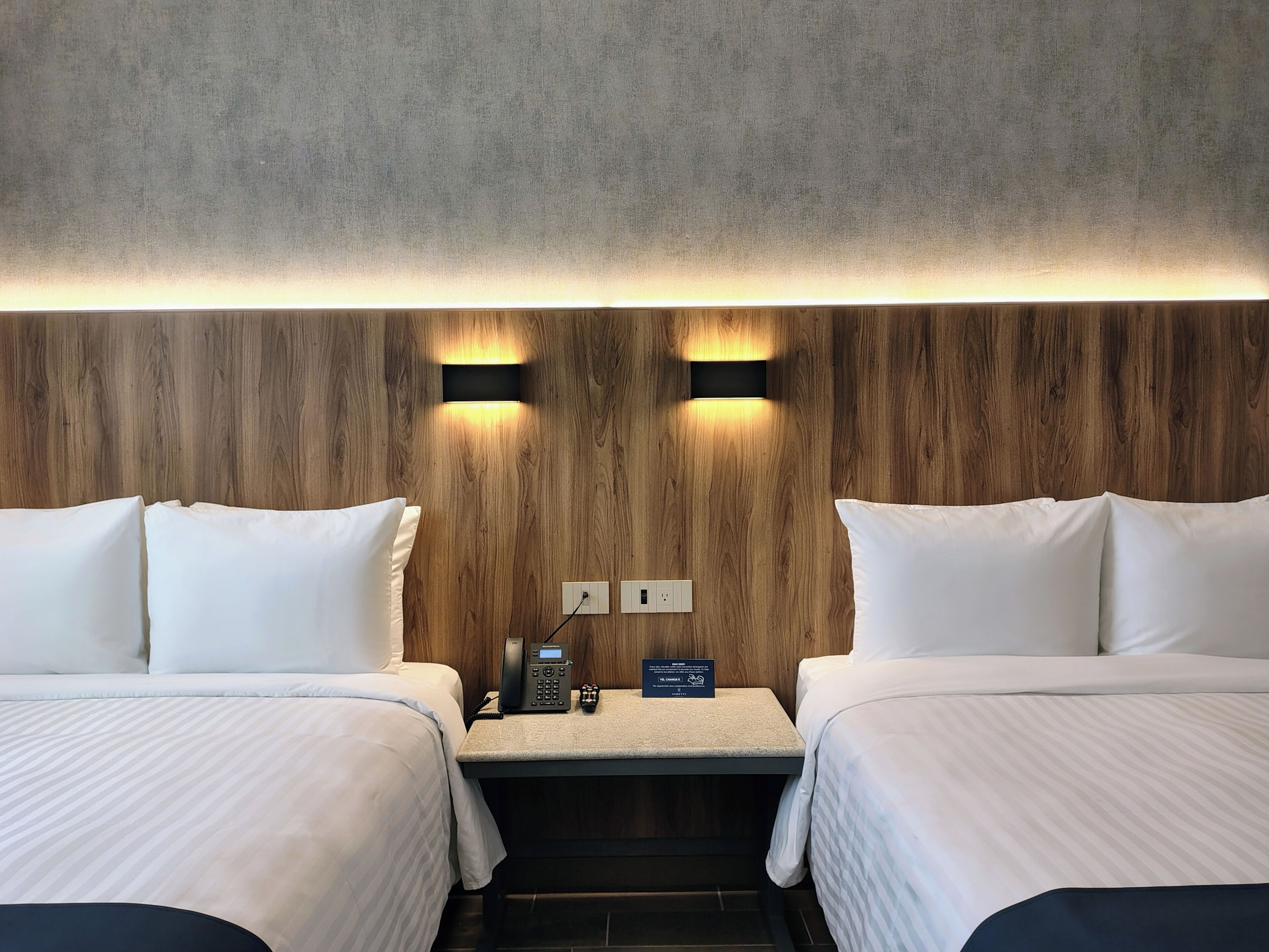 Habitación de hotel con dos camas, una mesita con un teléfono y un letrero, y una pared con textura.