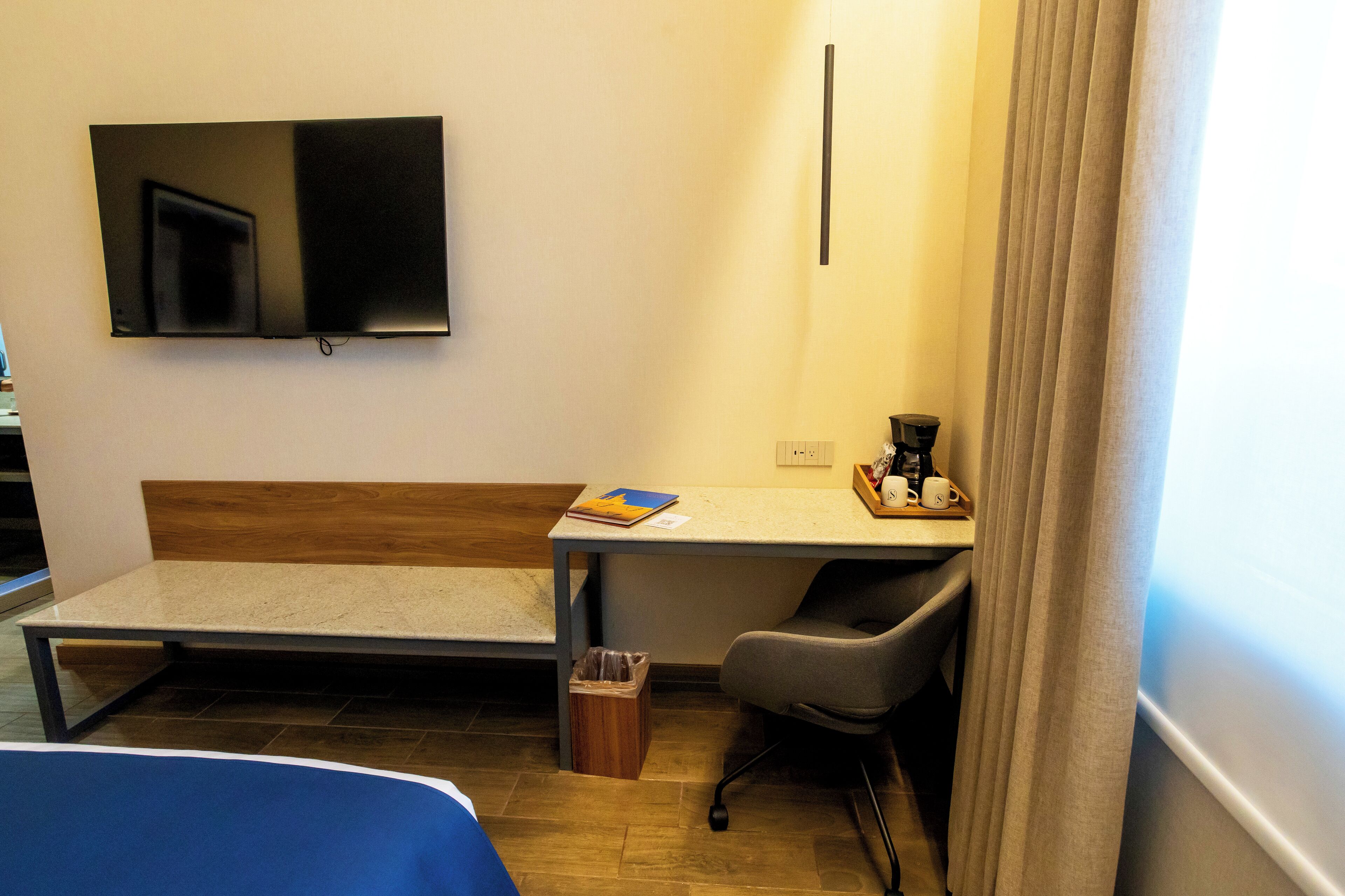 A hotel room with a bed, a desk with a chair, a TV mounted on the wall, and a window with curtains.