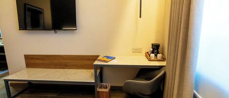 A hotel room with a bed, a desk with a chair, a TV mounted on the wall, and a window with curtains.