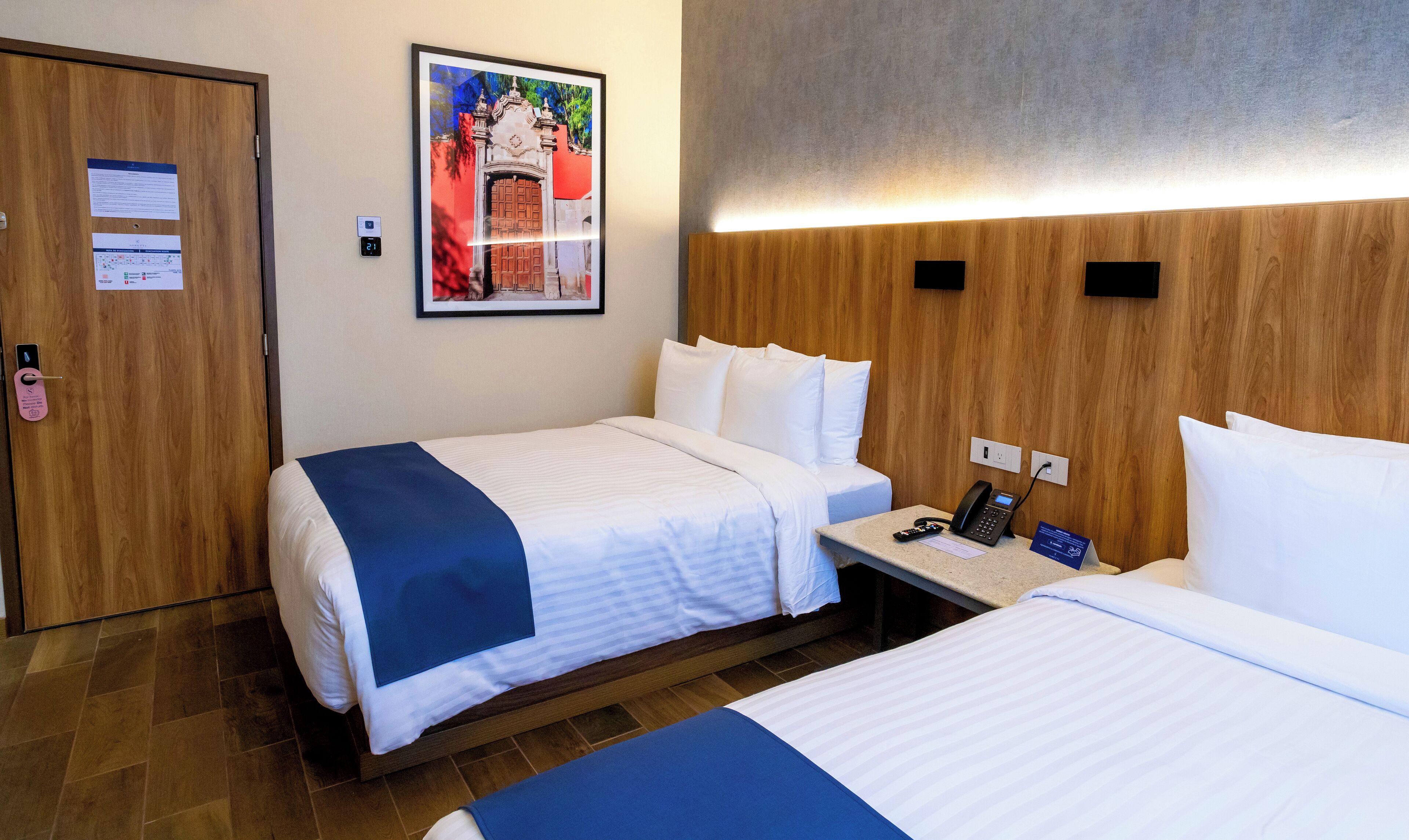 A hotel room with two beds, a wooden headboard, a nightstand with a phone, and a framed picture on the wall.