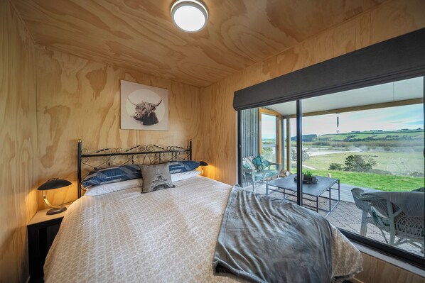 A bedroom with a large bed, a view of the countryside through a large window, and a small balcony area with a table and chairs. - Strathvale River Cottage - Winton Holiday Home (Winton)