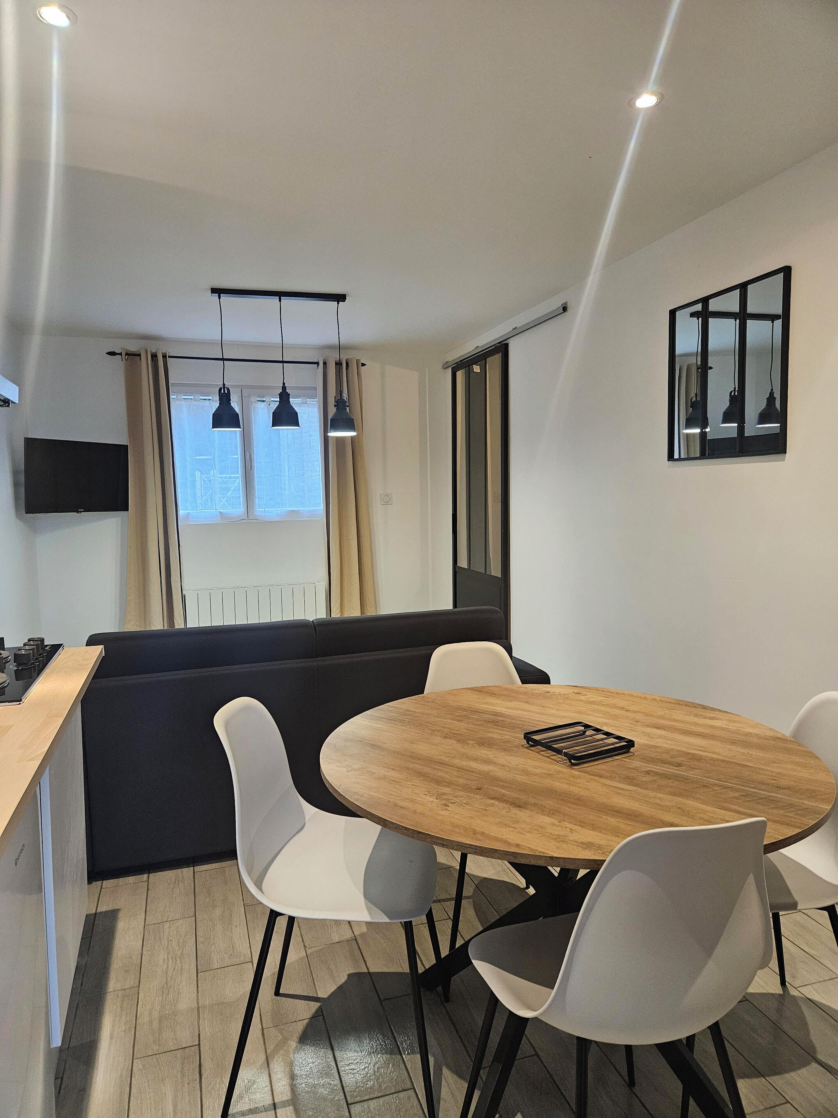 Een moderne keuken met een houten aanrecht, een zwarte kookplaat en een zwarte bank. Er staat een ronde houten tafel met vier witte stoelen, een zwarte hanglamp en een spiegel met een zwart frame aan de muur.