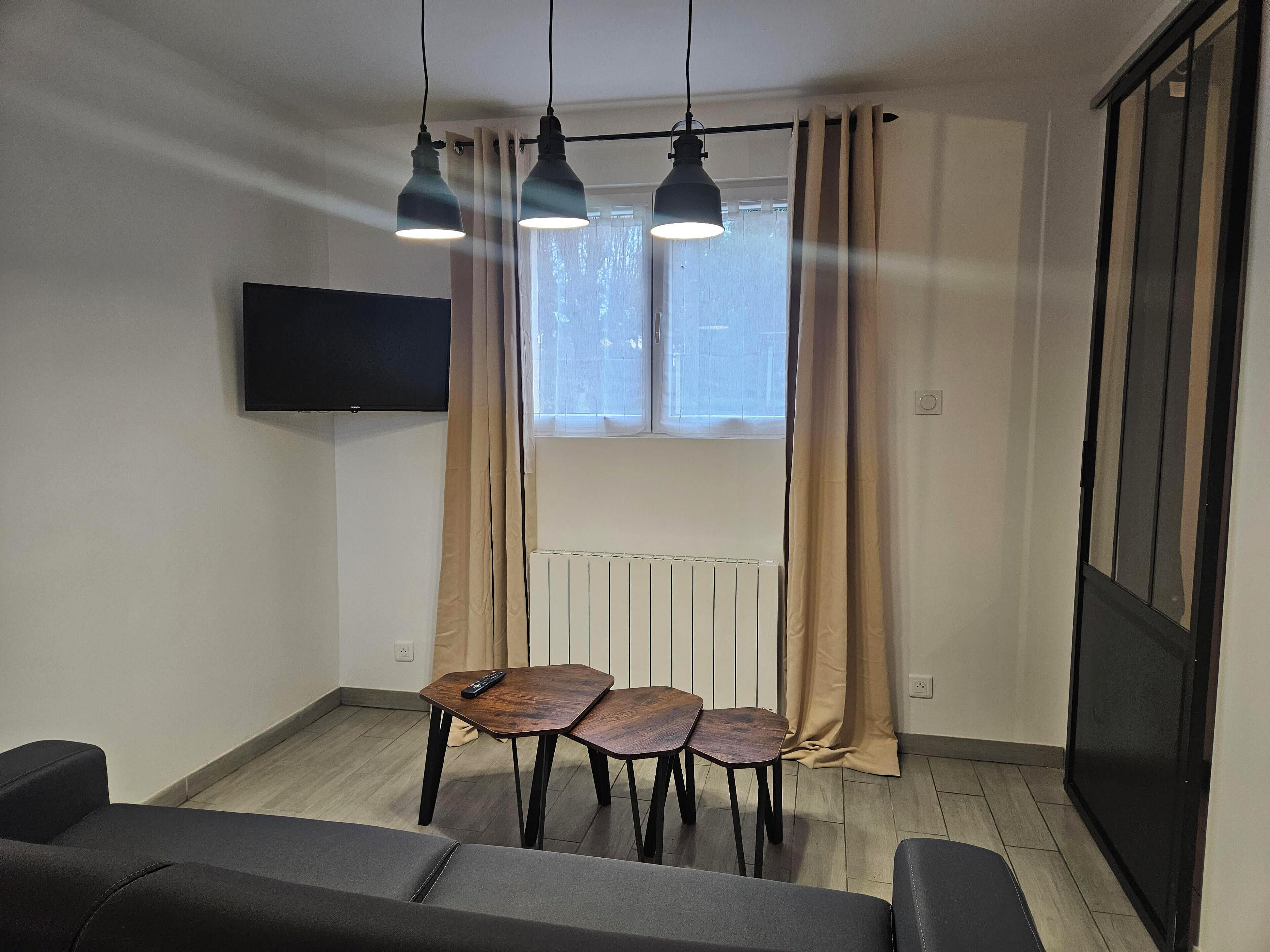 Ein modernes Wohnzimmer mit an der Wand montiertem Fernseher, einem Esstisch mit zwei Stühlen und einem Heizkörper unter einem mit Vorhängen drapierten Fenster.