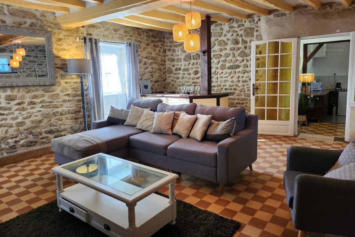 Ein gemütliches Wohnzimmer mit Steinwand, kariertem Boden, einer Couch mit Kissen, einem Glastisch und einer Holzbalkendecke.