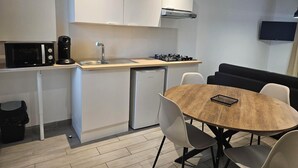 A compact kitchen with a microwave, coffee maker, sink, and stove. There is a small refrigerator, a dining table with chairs, and a black couch. A flat-screen TV is mounted on the wall. - Le Clos Loret (Saint-Germain-du-Corbéis)