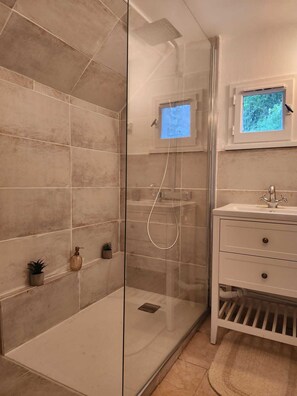 Ein modernes Badezimmer mit einer gläsernen Duschkabine, einem kleinen Fenster mit Ausblick und einem weißen Waschtisch mit Schubladen.