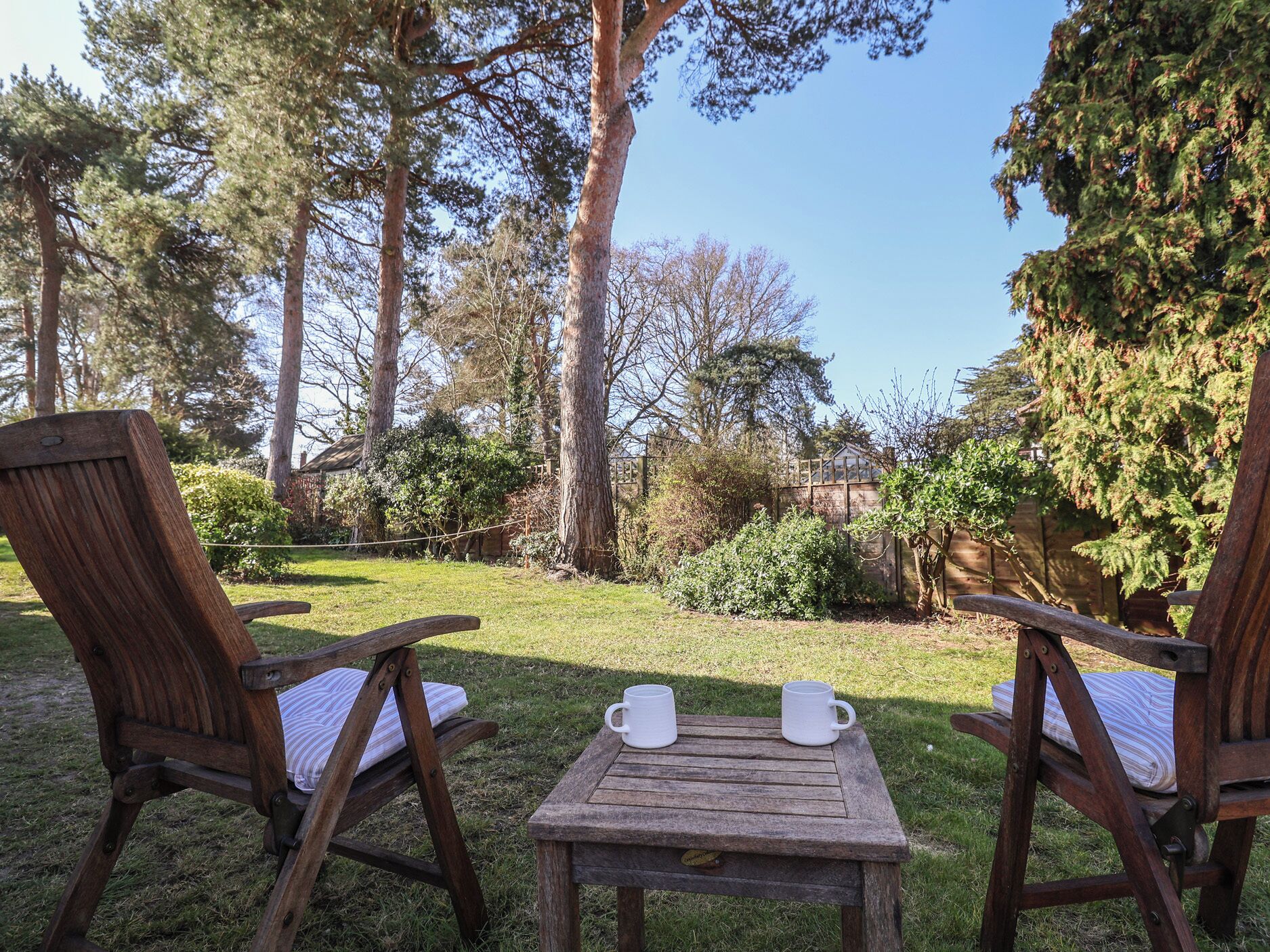 A wooden deck with two chairs and a small table, surrounded by a garden with trees and shrubs.