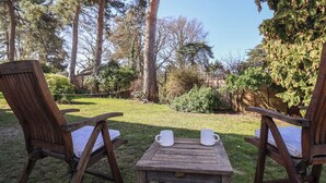 A wooden deck with two chairs and a small table, surrounded by a garden with trees and shrubs. - West End (Woodbridge)