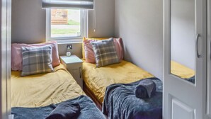 A room with two beds, each with a pillow and a blanket. There is a window with blinds, a small table with a lamp, and a door with a picture on it.