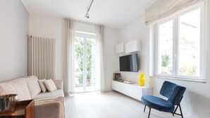 Een moderne woonkamer met een bank, een blauwe fauteuil, een televisie en een raam met uitzicht op groen.
