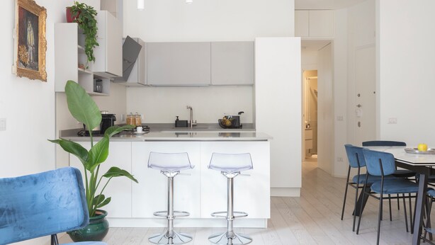 Een moderne keuken met witte kastjes, twee barkrukken, een eettafel met stoelen en een potplant.