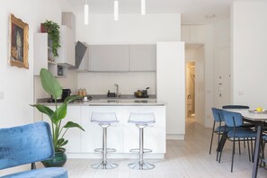 Une cuisine moderne avec des meubles blancs, deux tabourets de bar, une table à manger avec des chaises et une plante en pot.