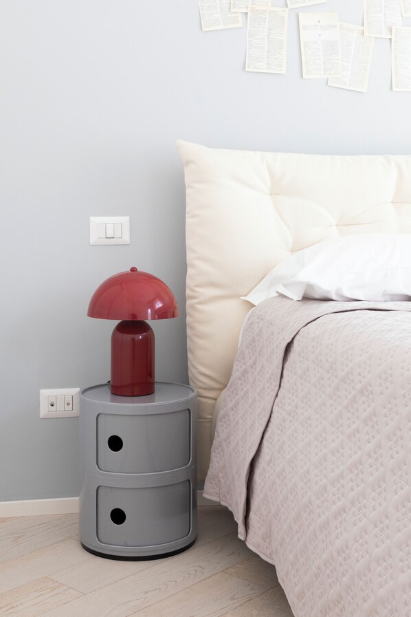 Un lit bien fait, avec une couette et des oreillers de couleur beige, une table de chevet grise sur laquelle se trouve une lampe rouge, et une prise électrique murale.