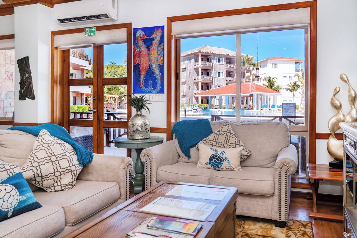 A hotel lobby with a beige sofa, a coffee table with magazines, a painting of seahorses, and a view of a building with a red roof.
