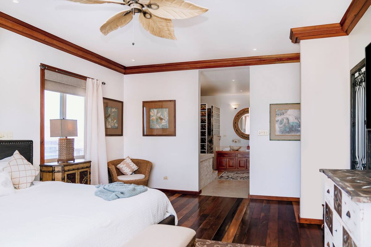A spacious bedroom with a large bed, a ceiling fan, wooden flooring, and a view into the bathroom.