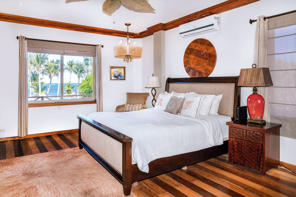 A bedroom with a wooden bed, a chandelier, a nightstand, and a view of palm trees through a window.