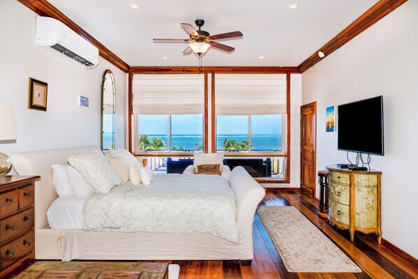 A bedroom with a large bed, a ceiling fan, a TV, and a view of the ocean.