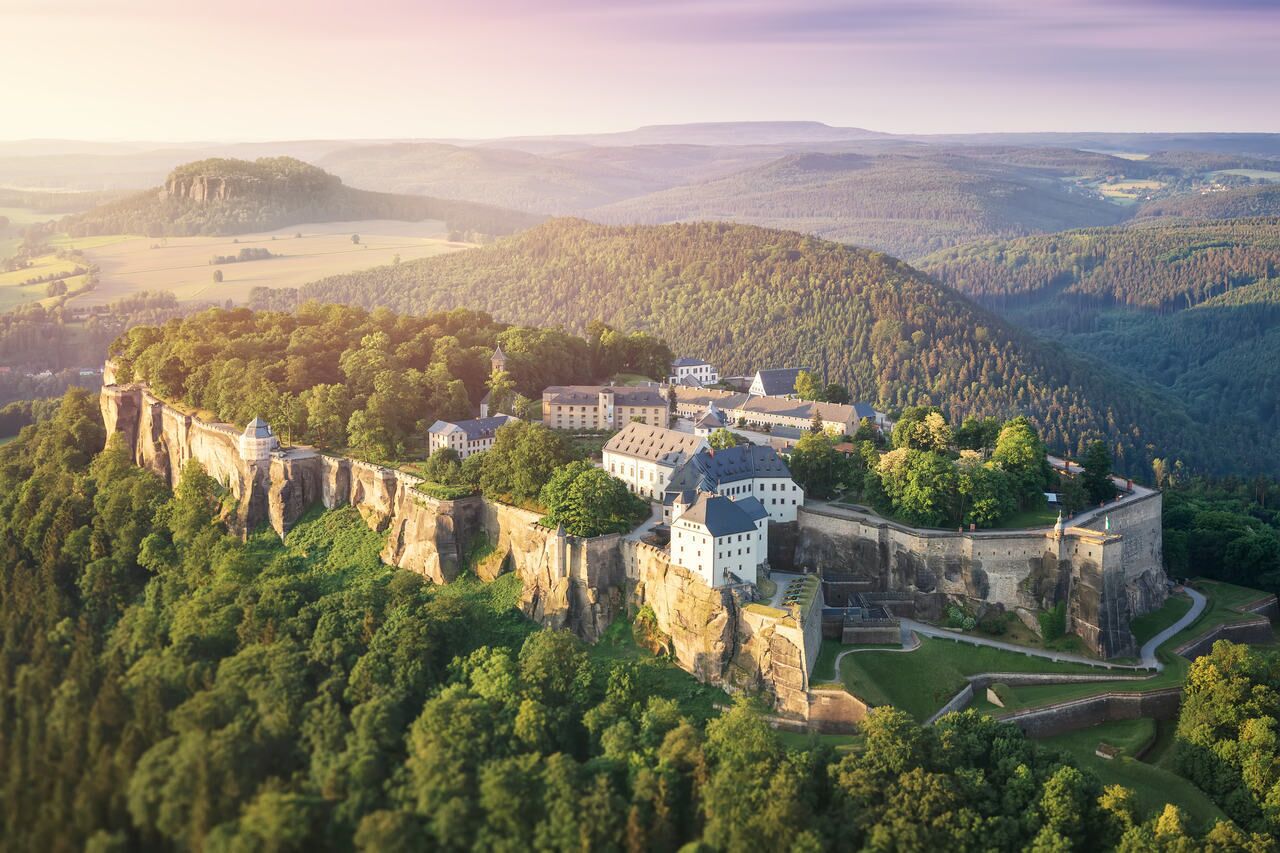 A castle perched on a cliff with multiple buildings, surrounded by dense forest and rolling hills.