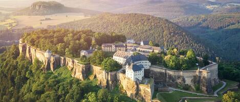 A castle perched on a cliff with multiple buildings, surrounded by dense forest and rolling hills.