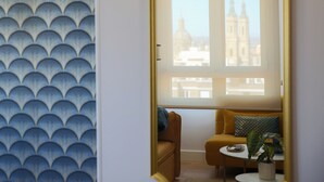 Ein Zimmer mit Wandmuster, einer Couch mit Kissen, einem kleinen Tisch mit Pflanze und einem Fenster mit Blick auf Gebäude.