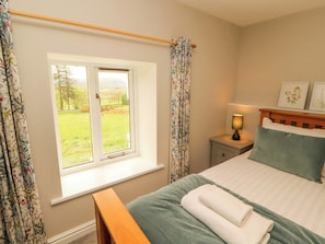 A bedroom with a bed, a bedside table with a lamp, a window with curtains, and a view of a grassy field. - Swallow's Nest (Keswick)
