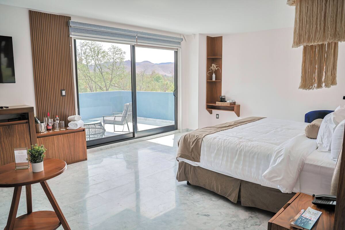 A hotel room with a bed, a desk, a chair, a small table with a plant, a shelf with decorative items, and a view of the outdoors through a sliding door.