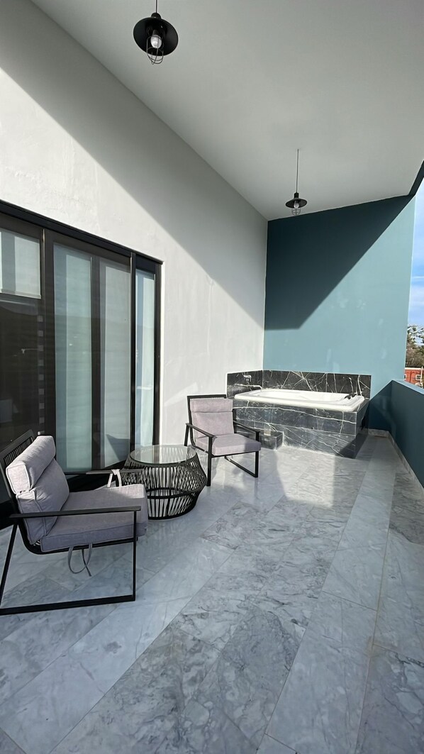 A modern balcony with a glass enclosure, a seating area with a sofa and chairs, a marble-tiled floor, and a hot tub.