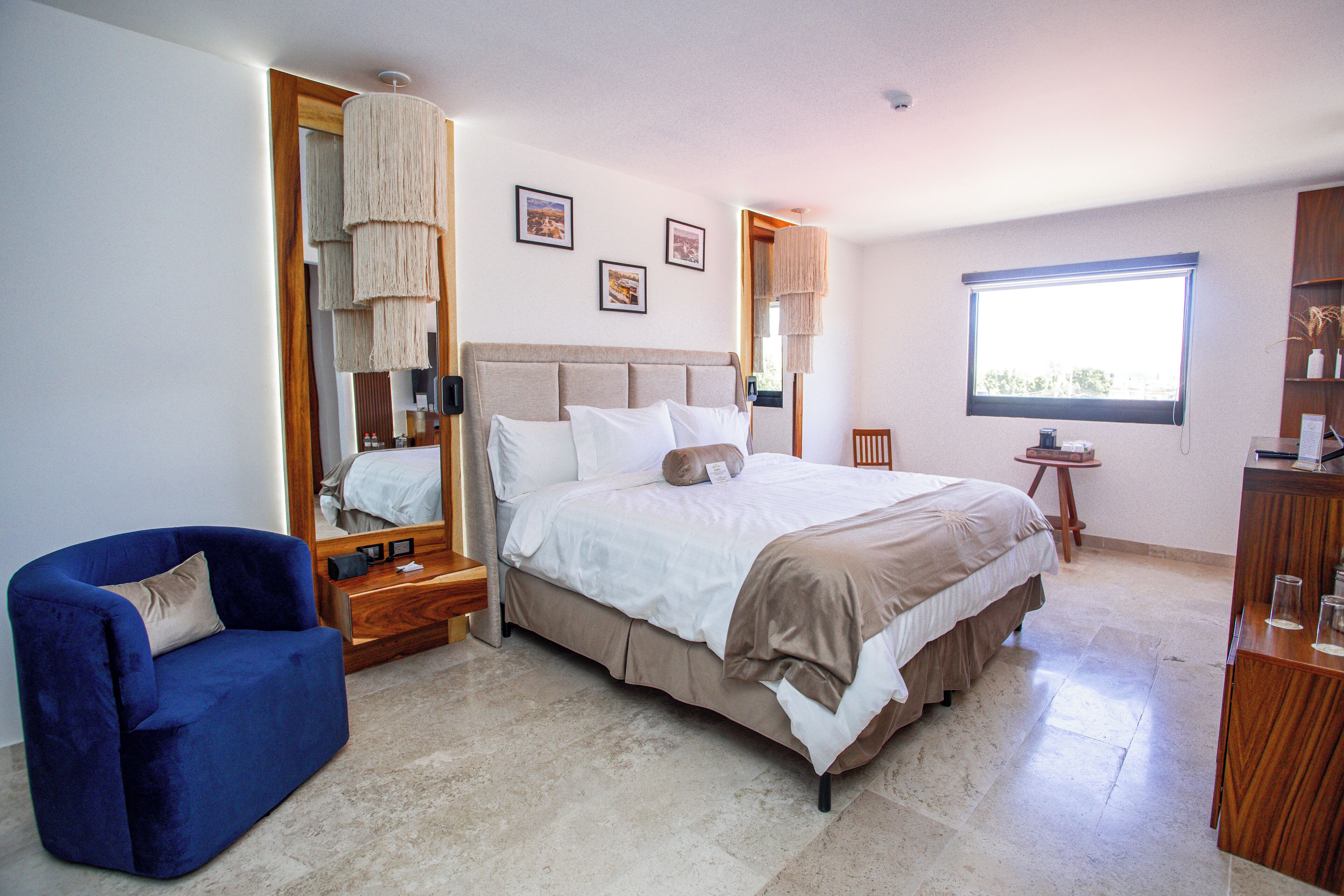 Un dormitorio con una cama, un sillón azul, una mesita de noche de madera, un espejo y una ventana.