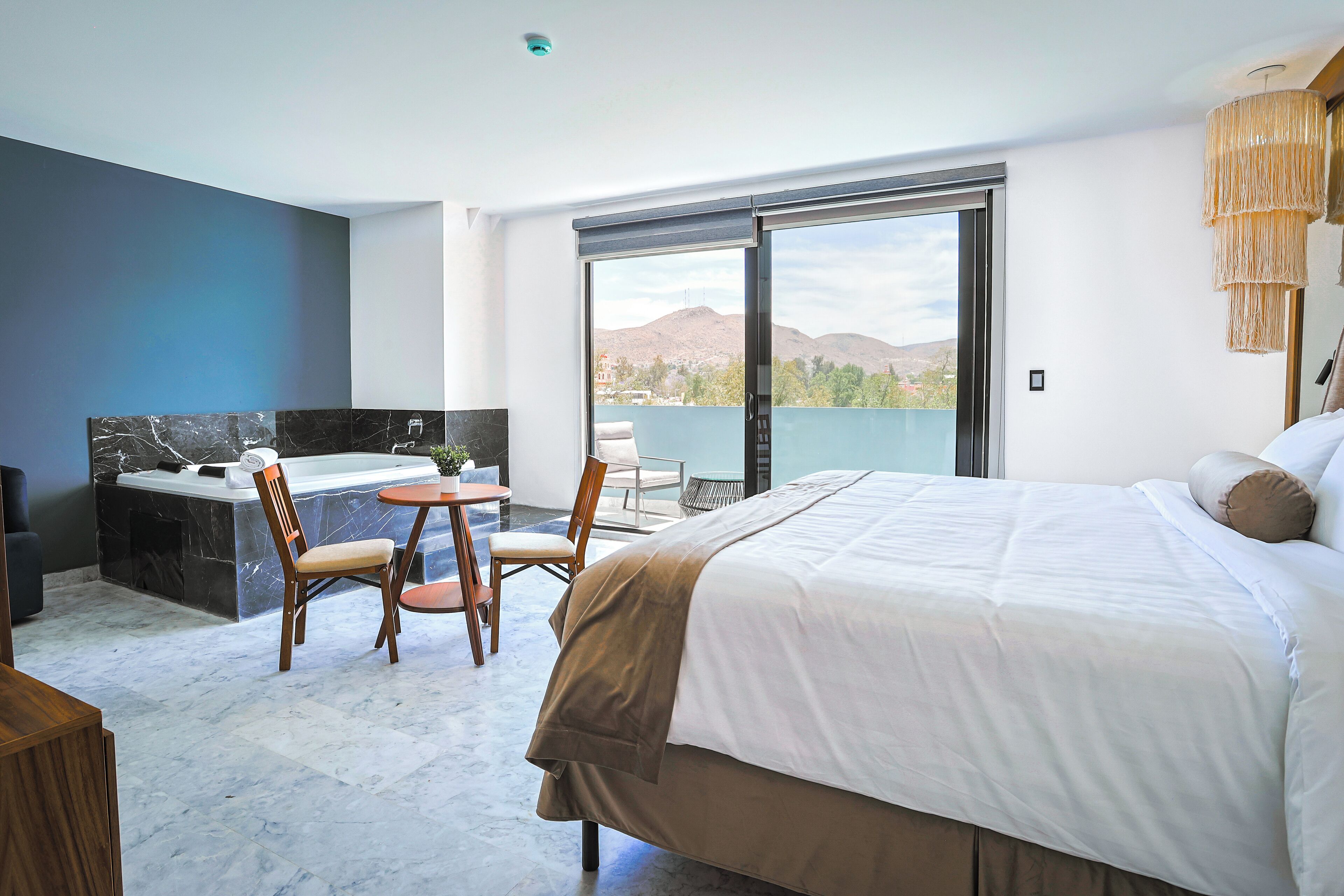 A modern hotel room with a large bed, a sitting area with a table and chairs, a bathtub, and a balcony with a view of the mountains.